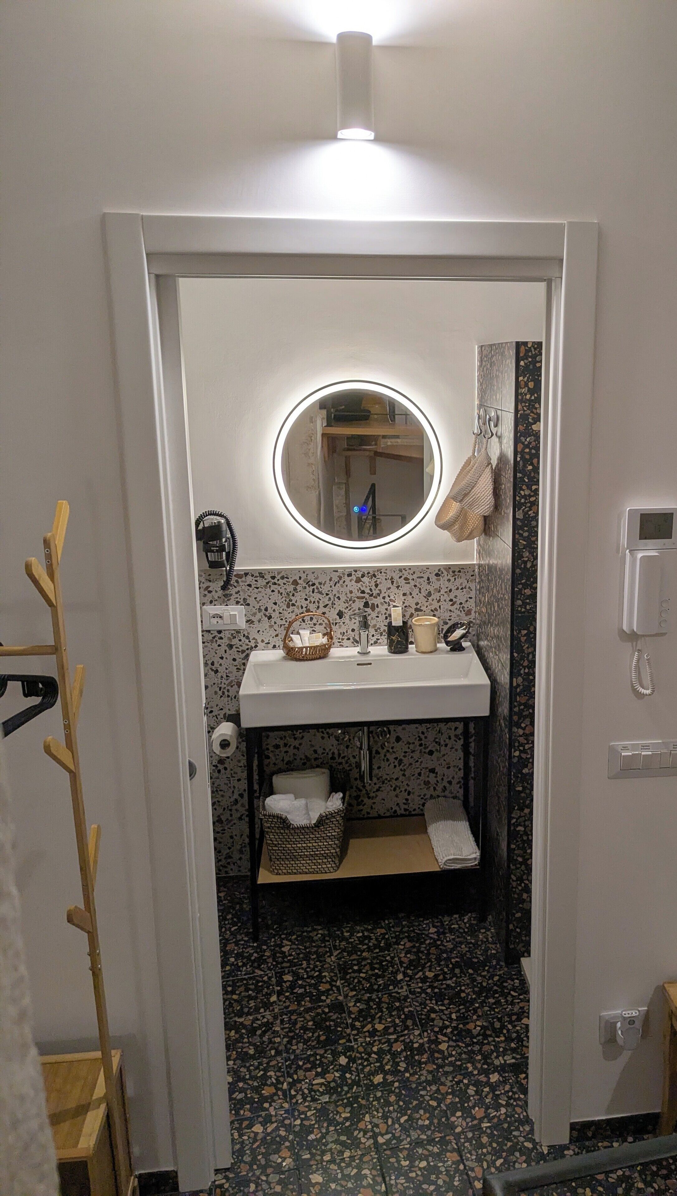 Een badkamer met een witte wastafel, een ronde spiegel, een handdoekhouder, een telefoon en een klein plankje met toiletartikelen.