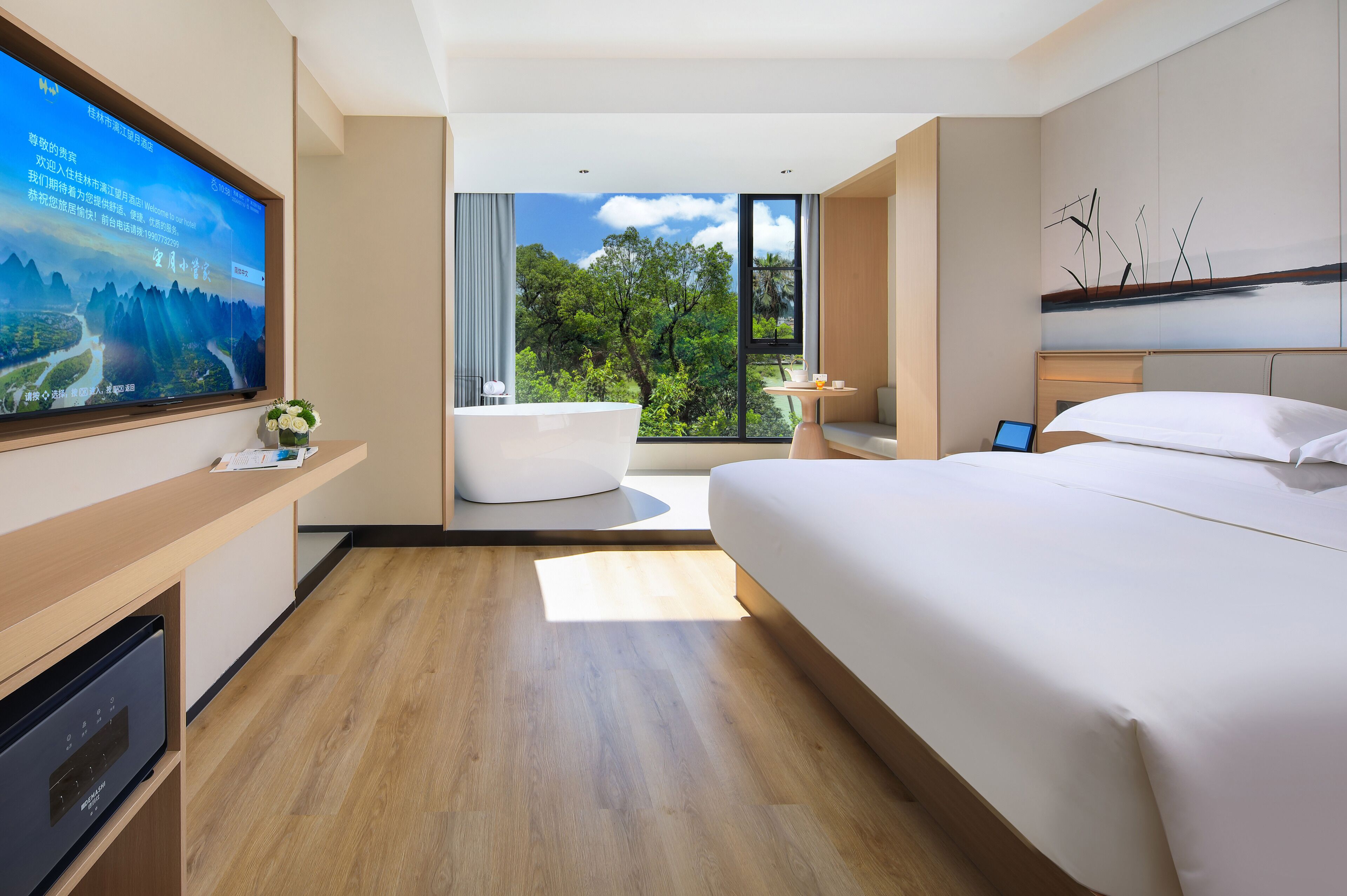 A modern hotel room with a large bed, a bathtub, a TV, and a view of greenery.