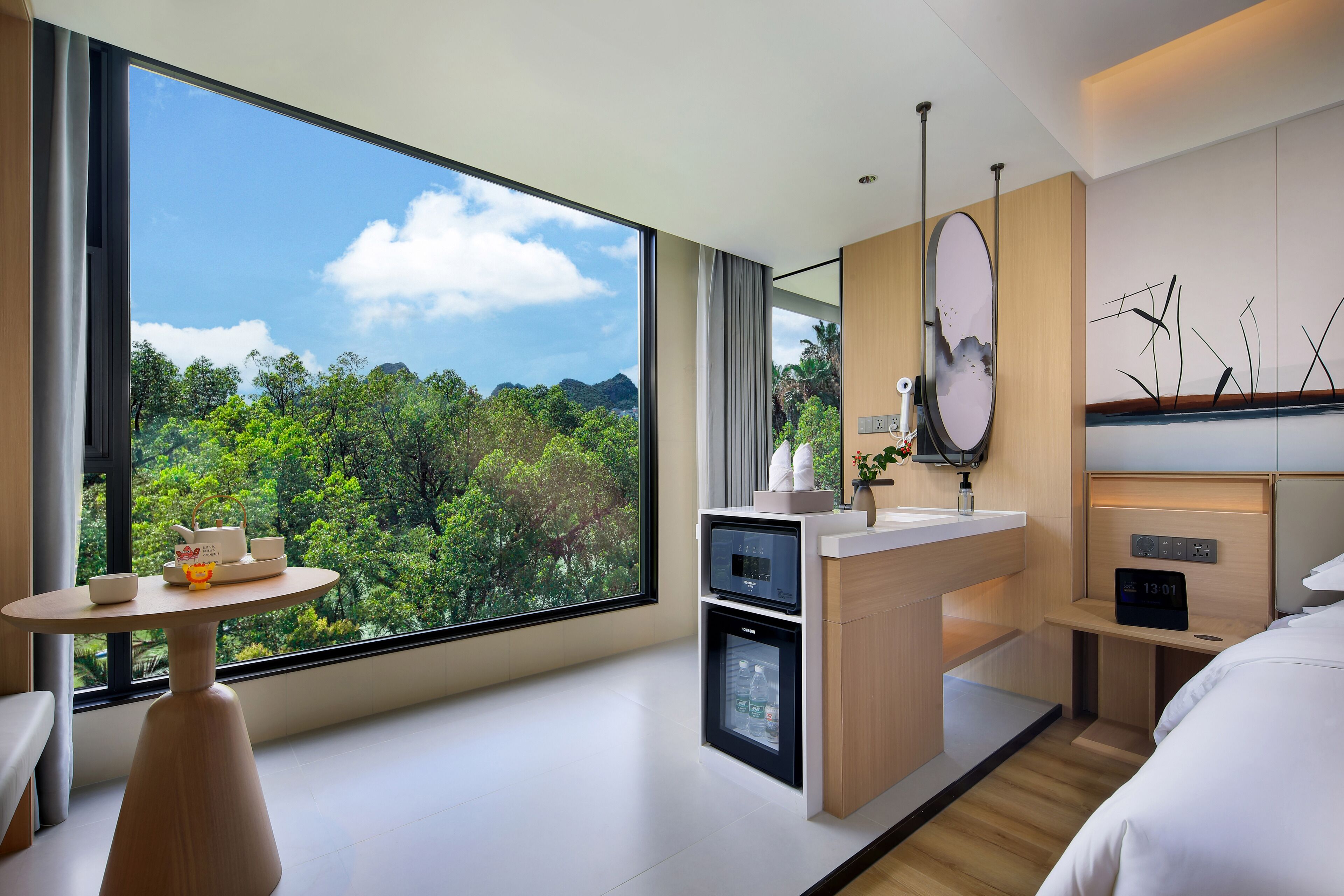 A modern hotel room with a large window, a compact kitchenette, a bedside table, and a bed.