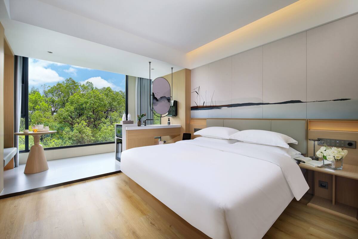 Una habitación de hotel moderna con una cama grande, una mesita de noche con un florero, un escritorio con una silla y un ventanal que ofrece vistas a árboles y al cielo.
