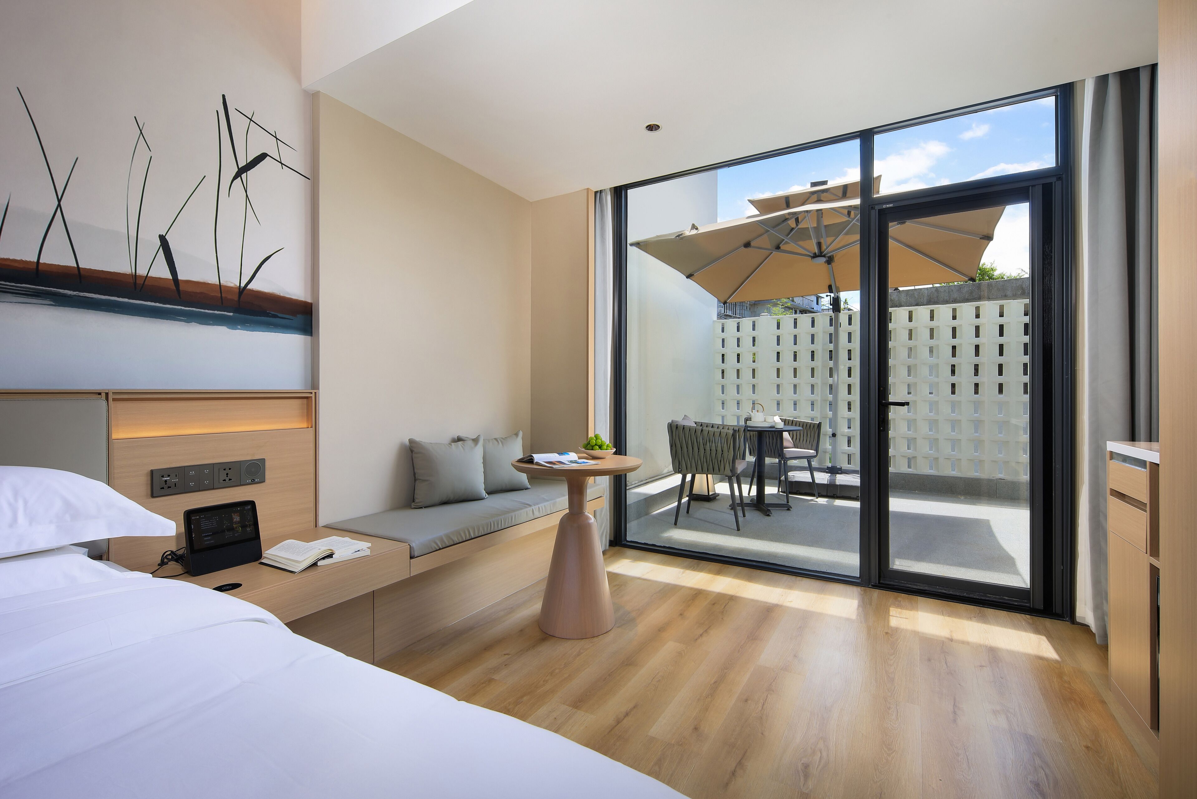 Quarto de hotel moderno com cama, mesa de cabeceira e vista do pátio exterior através de amplas portas de vidro deslizantes.