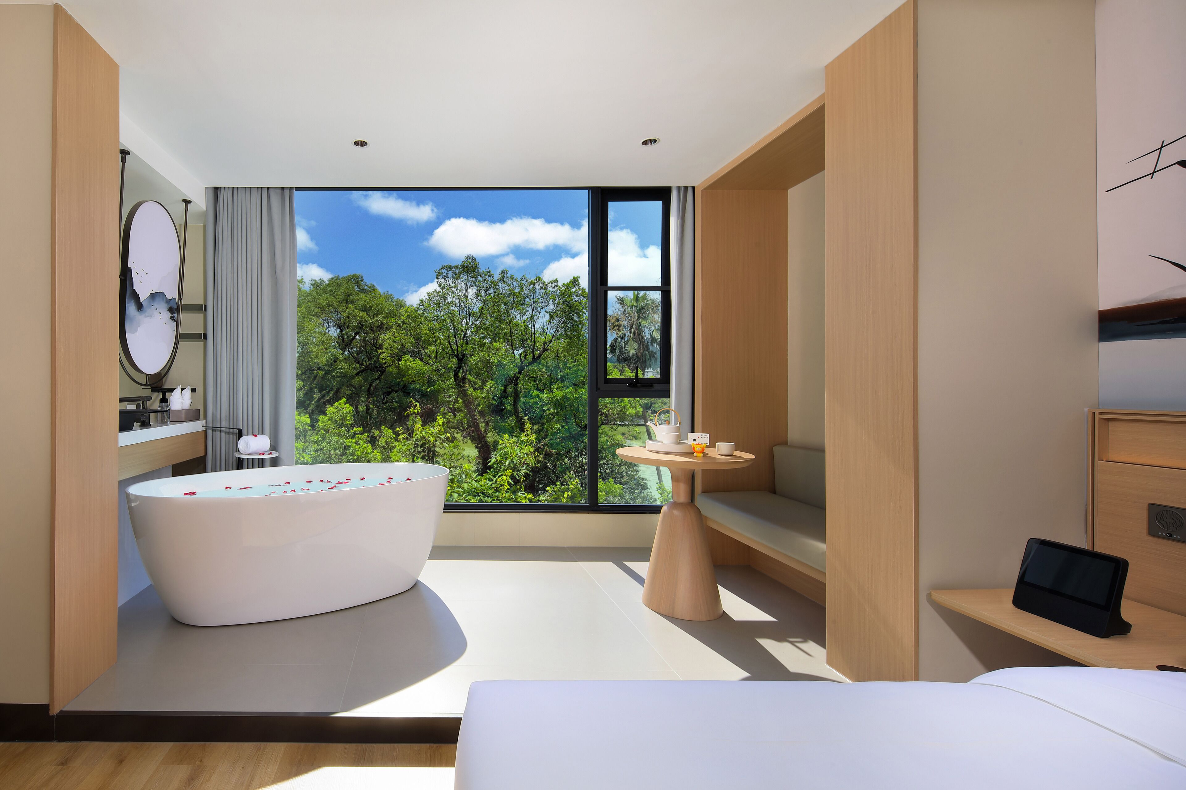 A modern hotel room with a large bathtub, a bedside table with a tablet, and a view of lush greenery through large windows.