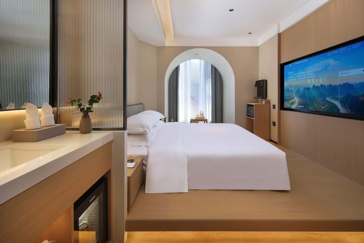 Una habitación de hotel moderna con una cama grande, un televisor de pantalla plana, una mesita de noche con un jarrón de flores y un ventanal con cortinas.