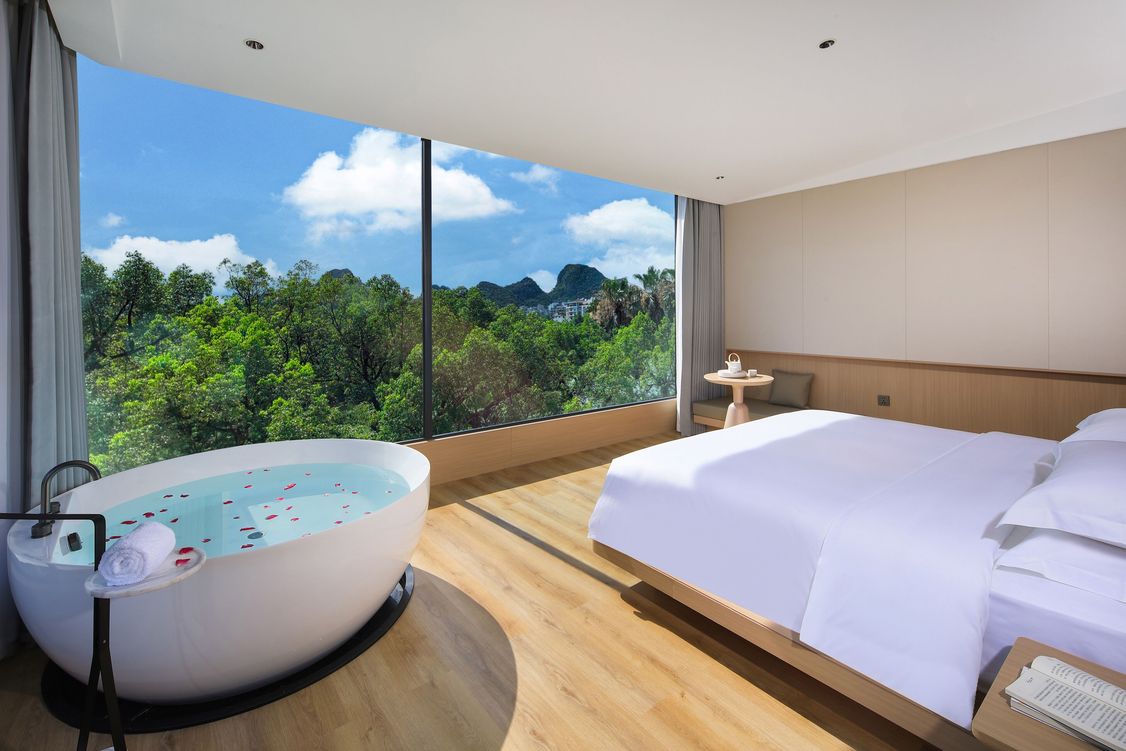 Una habitación de hotel moderna con una cama grande, una bañera con pétalos de rosa y vistas a follaje exuberante y montañas.