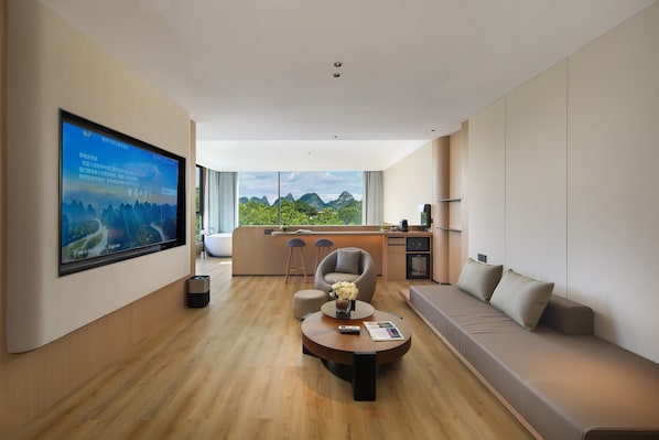 Ein modernes Wohnzimmer mit einem großen Flachbildfernseher, einem runden Couchtisch, einer Couch und Blick auf Grünflächen durch große Fenster.