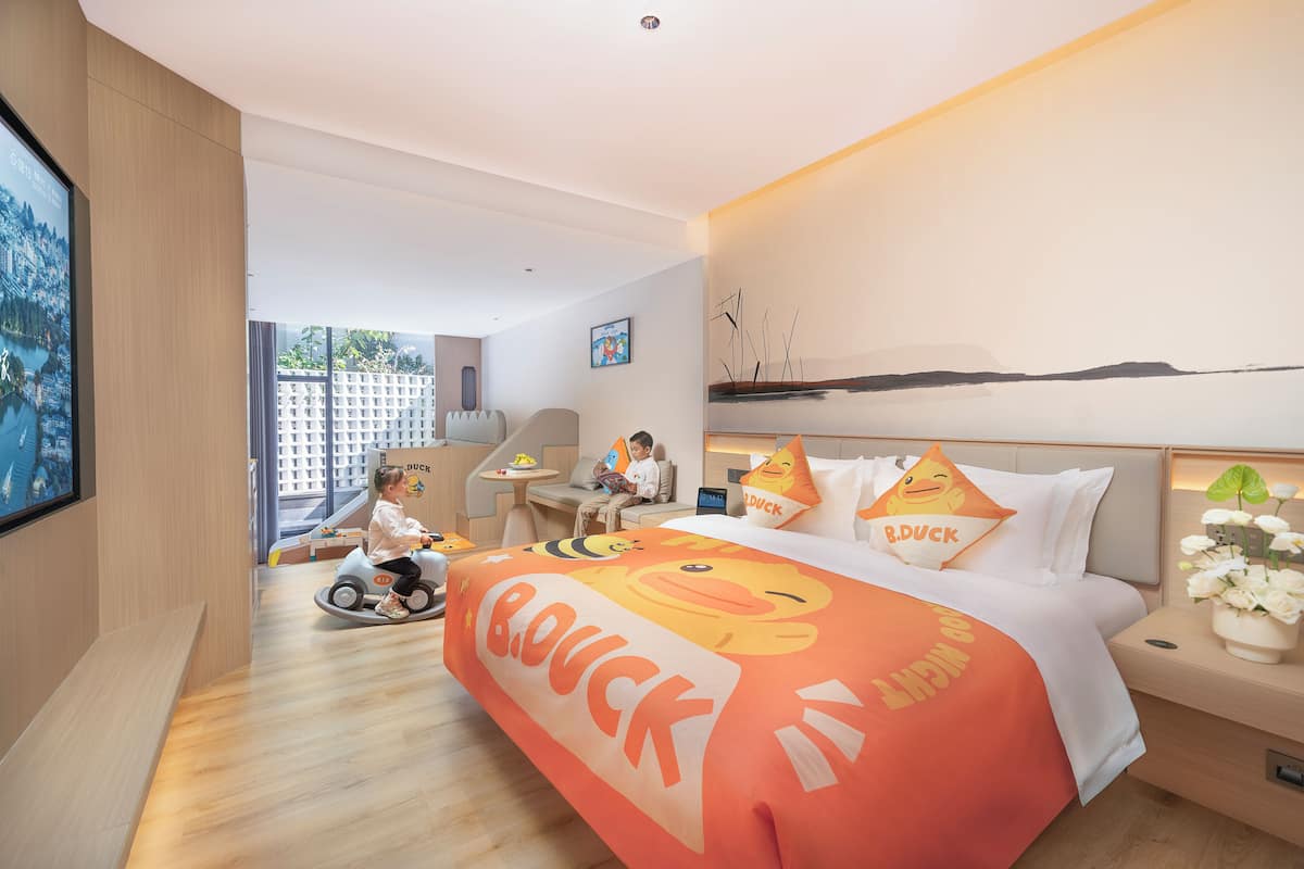 Habitación de hotel con una cama decorada con motivos de patos, un juguete para que juegue un niño y vistas a un paisaje urbano a través de grandes ventanales.