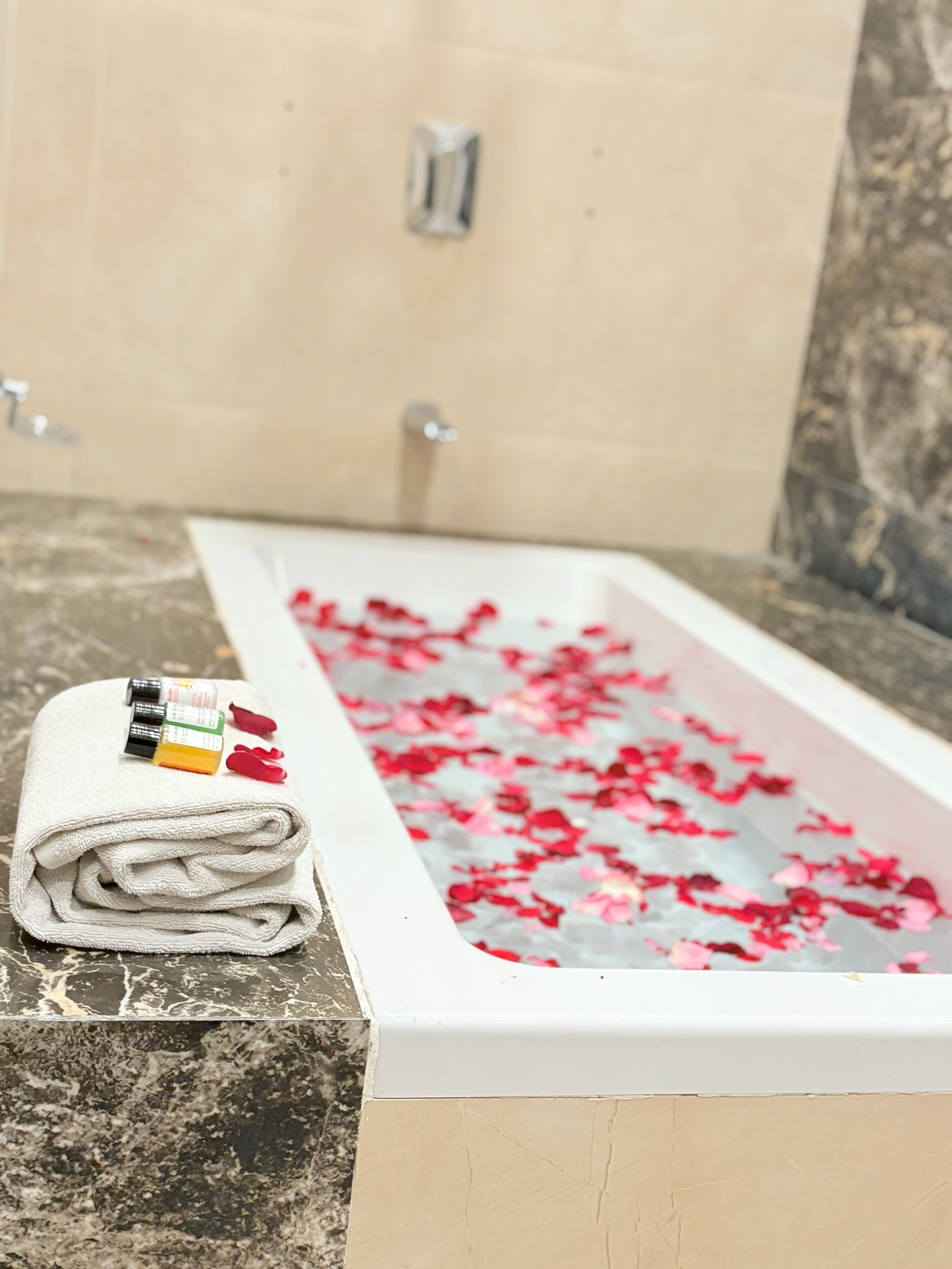 A bathroom with a bathtub filled with rose petals, a folded towel, and a bottle of lotion on the counter.