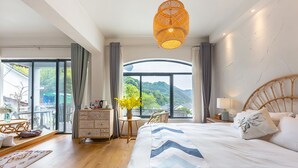 A bedroom with a large bed, a wooden headboard, a bedside table with a lamp, a window with a view of greenery, and a balcony with a table and chairs.