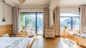 A spacious bedroom with a large bed, a wooden dresser, a small table, and a chair. There are large windows with curtains, a chandelier, and a view of a balcony with outdoor furniture.