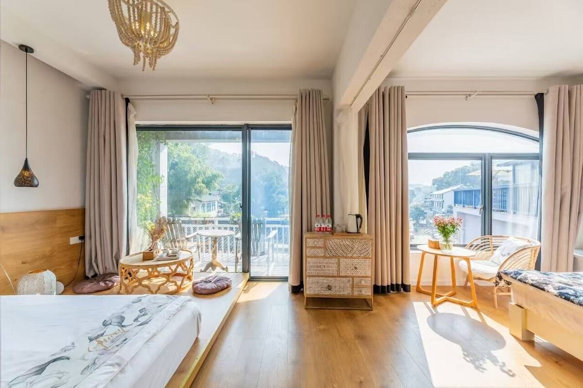 Une chambre spacieuse avec un grand lit, une commode en bois, une petite table et une chaise. De grandes fenêtres sont agrémentées de rideaux, une suspension orne le plafond, et la vue donne sur un balcon aménagé avec du mobilier d’extérieur.