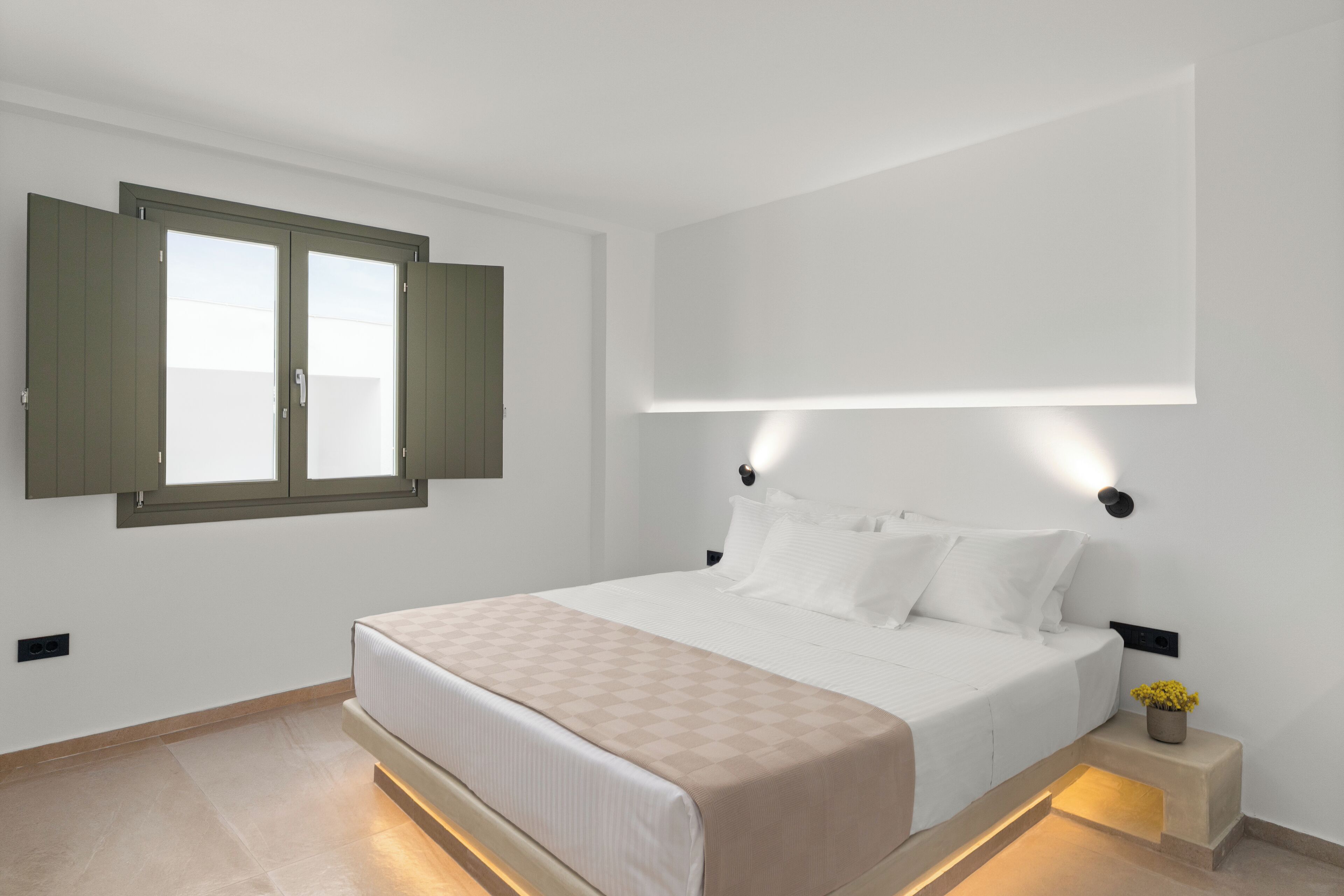 A bedroom with a bed, a window with open shutters, a bedside table with a lamp, and a wall-mounted light fixture.