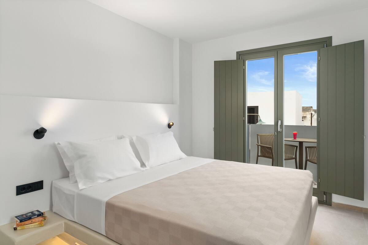 A modern hotel room with a large bed, a bedside table with books, a small table with chairs, and a balcony with a table and chairs.