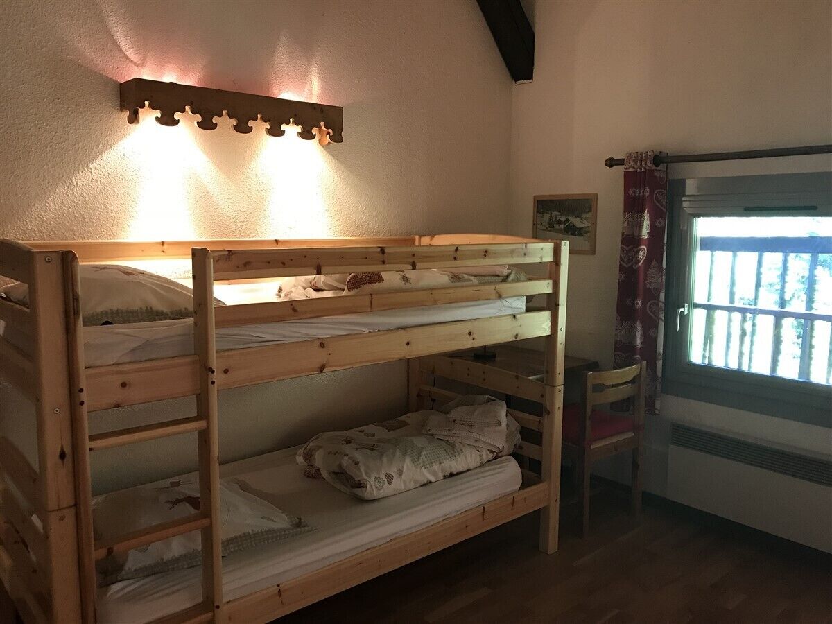 A bunk bed with a wooden frame, a wall-mounted light fixture, a window with a view, and a chair.