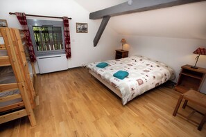 A bedroom with a bunk bed, a single bed with a patterned bedspread, a window with curtains, a bedside table with a lamp, and a small wooden chair.