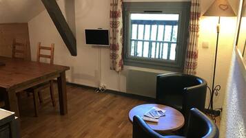 Een kamer met een houten eettafel, twee zwarte stoelen, een klein rond tafeltje met een afstandsbediening, een aan de muur bevestigde televisie, een raam met gordijnen en een lamp.