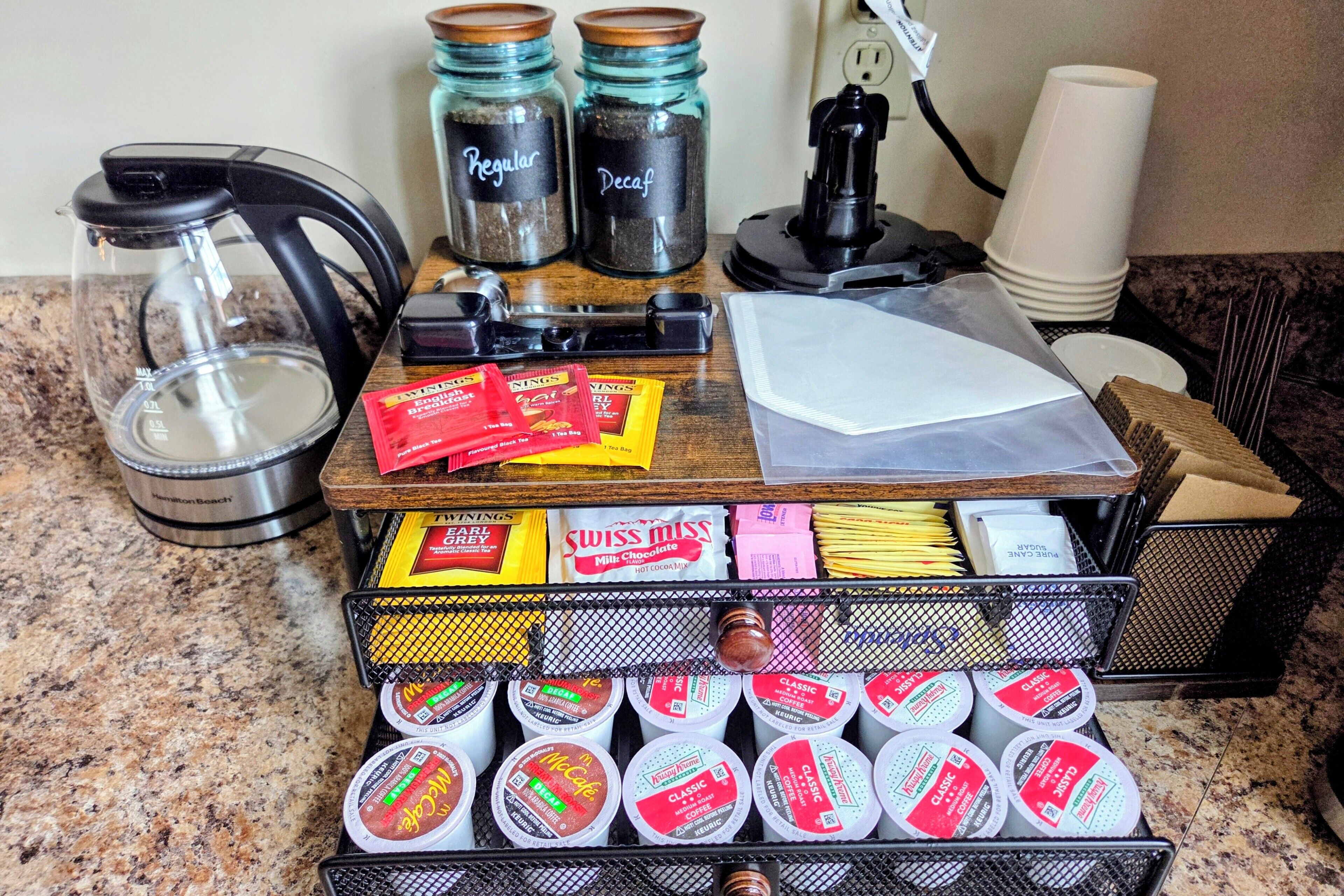 En köksbänk med diverse utrustning för att göra kaffe, inklusive en kaffebryggare, burkar märkta med ”vanligt kaffe” och ”kaffe utan koffein” samt ett fat med tepåsar och kaffekapslar.
