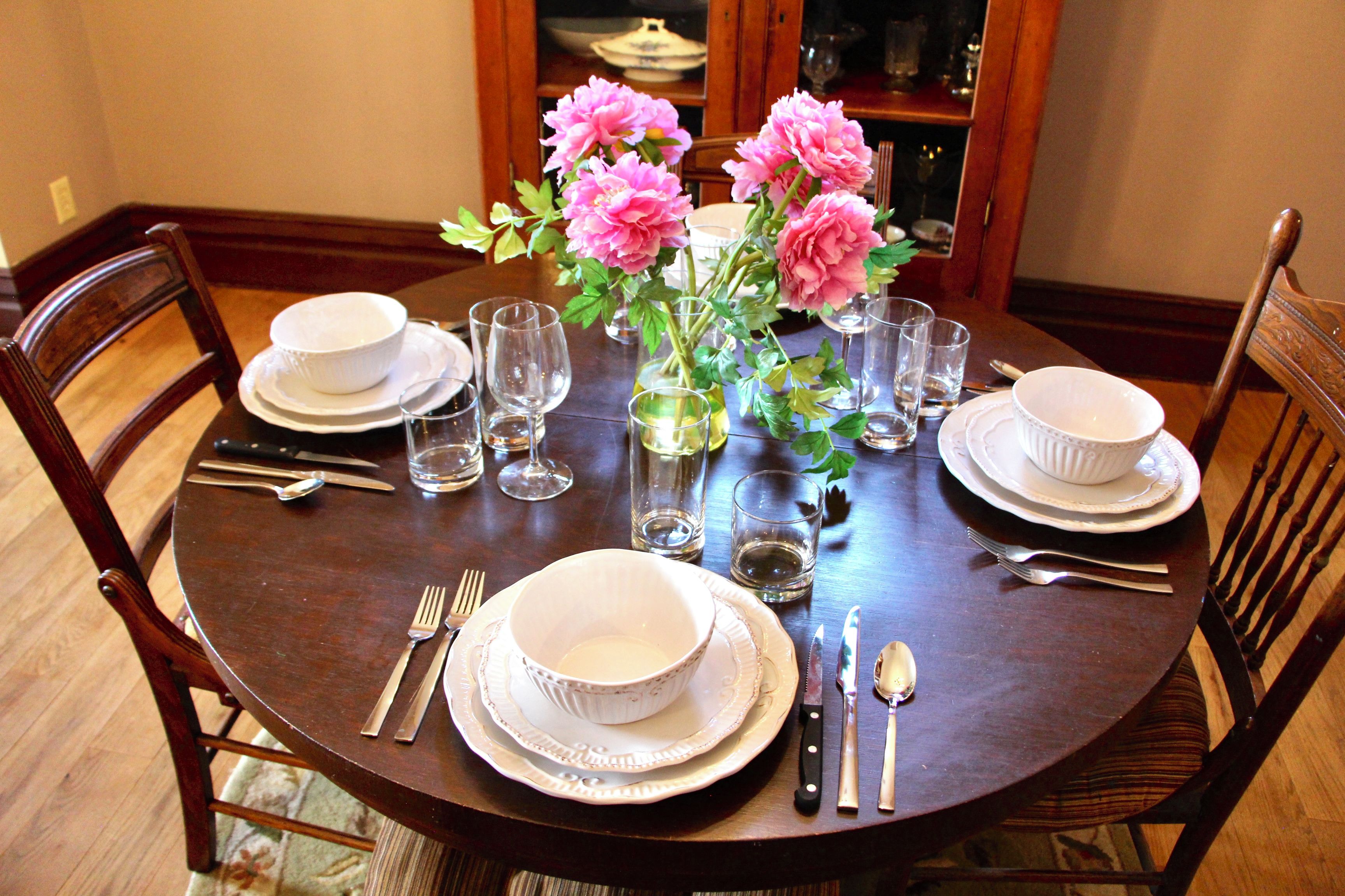 Una mesa de comedor dispuesta con platos, fuentes, cubiertos y vasos blancos, con un florero de flores rosadas en el centro.