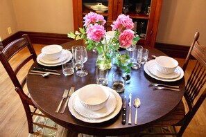 Une table à manger dressée avec de l’assiette blanche, des bols, des couverts et des verres, et un vase rempli de fleurs roses placé au centre.