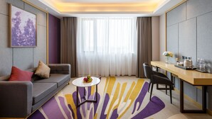 A modern hotel room with a grey sofa, a small round table with fruit, a dining area with a wooden table and chairs, and a large window with sheer curtains. - Lavande Hotel - Huizhou Boluo Shiwan (Huizhou)