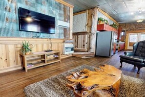 A living room with a wooden coffee table, a flat-screen TV mounted on a wall, a refrigerator, and a black leather sofa.