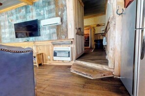 Un interior rústico con piso de madera, una zona de cocina con refrigerador y un televisor montado en la pared.