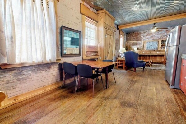 A rustic interior with a wooden dining table, chairs, a refrigerator, and a brick wall.