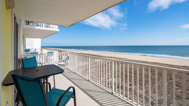 Ein Balkon mit Tisch und Stühlen, der Blick auf einen Strand mit Zaun und klarem Himmel gewährt.