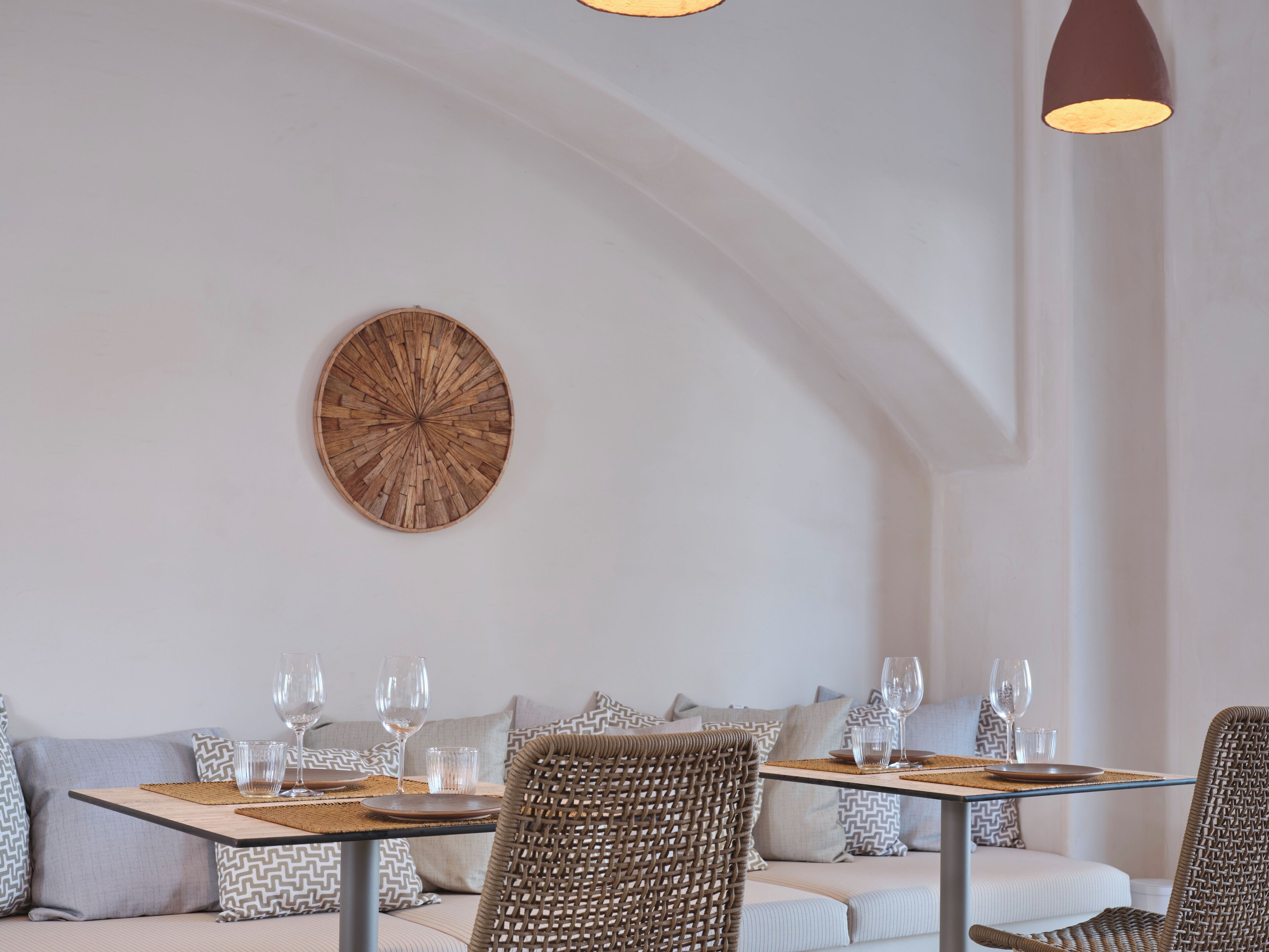 A dining area with a round wooden wall art piece, wicker chairs, and a table set for two with wine glasses.
