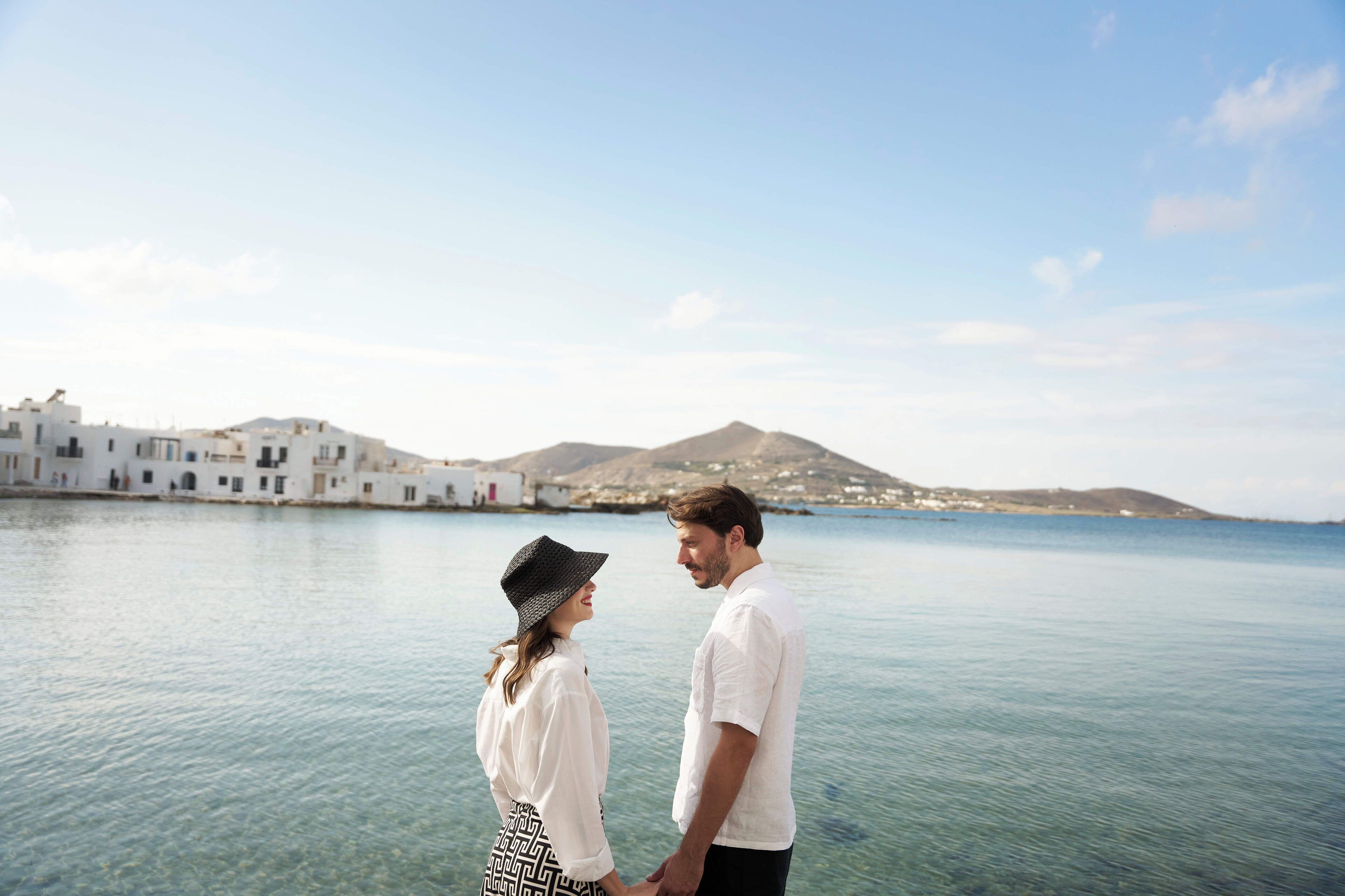 A couple holding hands by a body of water with white buildings in the background.