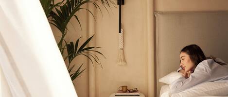 Une chambre à coucher avec un lit, une table de chevet sur laquelle se trouvent un livre et une tasse, une lampe suspendue et une plante en pot.