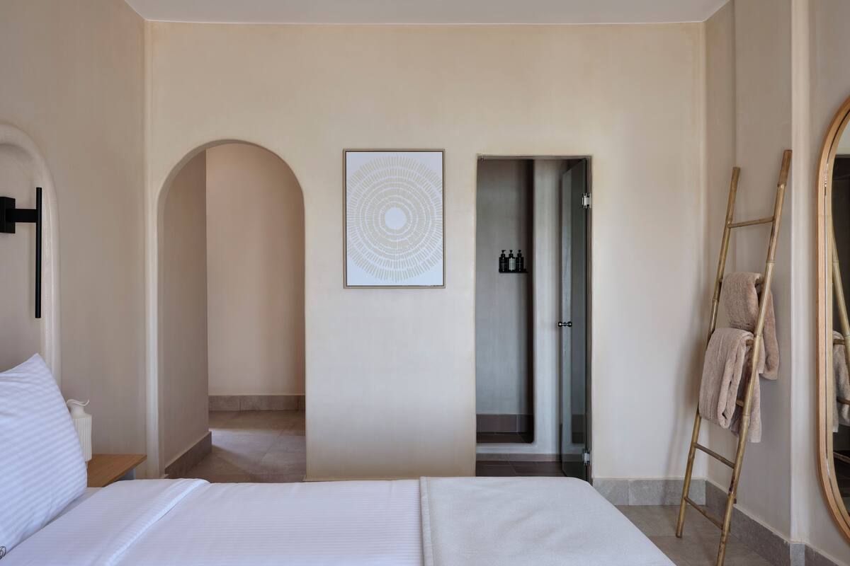 Quarto de hotel com uma cama grande, uma escada de madeira, uma obra de arte emoldurada e um banheiro visível através de um arco.