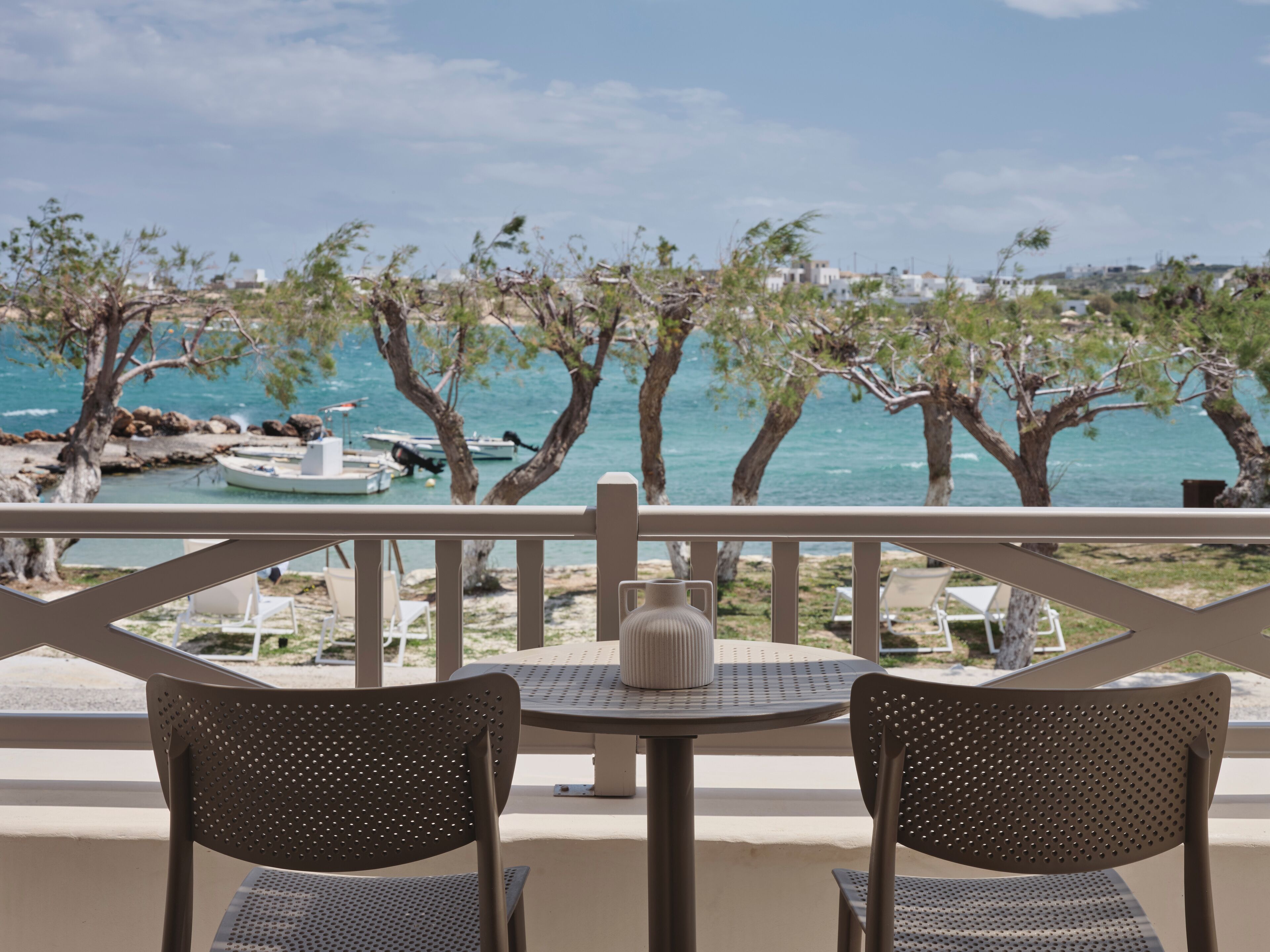 Un balcón con dos sillas y una mesa, con vista a una playa que tiene barcos y árboles.