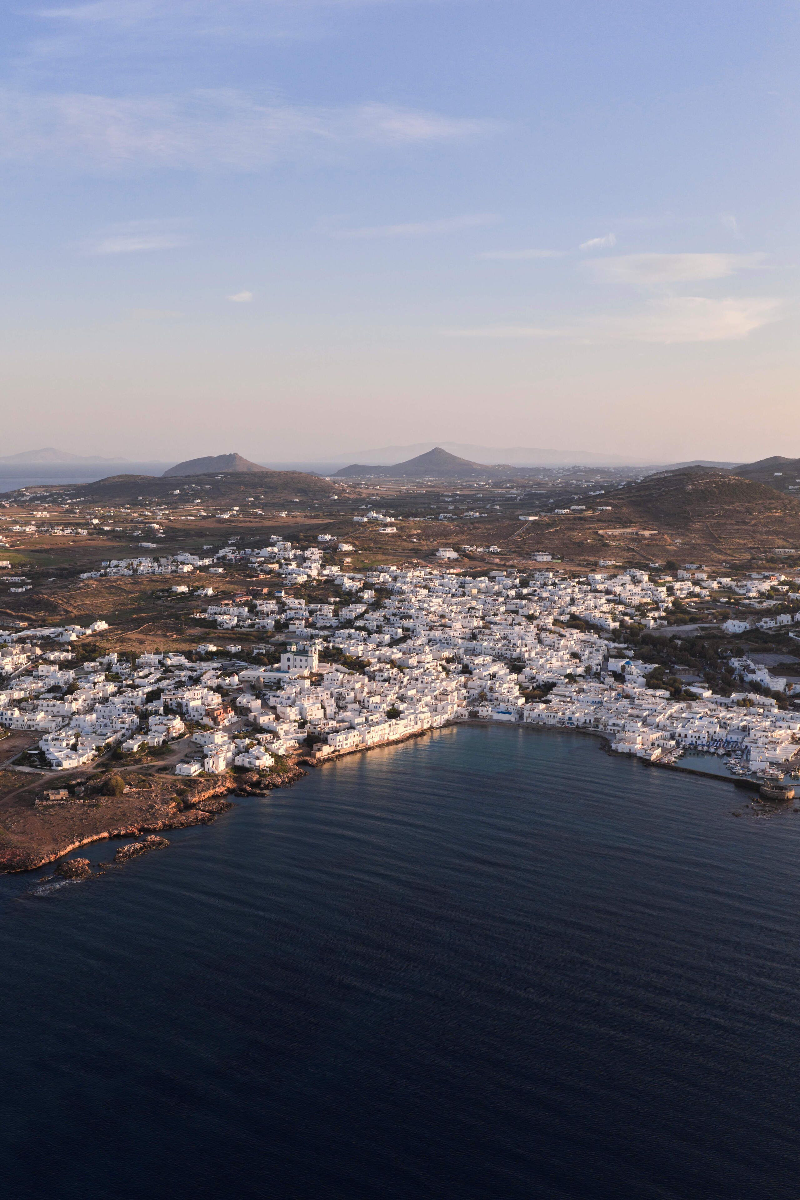 An aerial view of a coastal town with white buildings clustered along the shoreline, a clear sky, and distant hills.