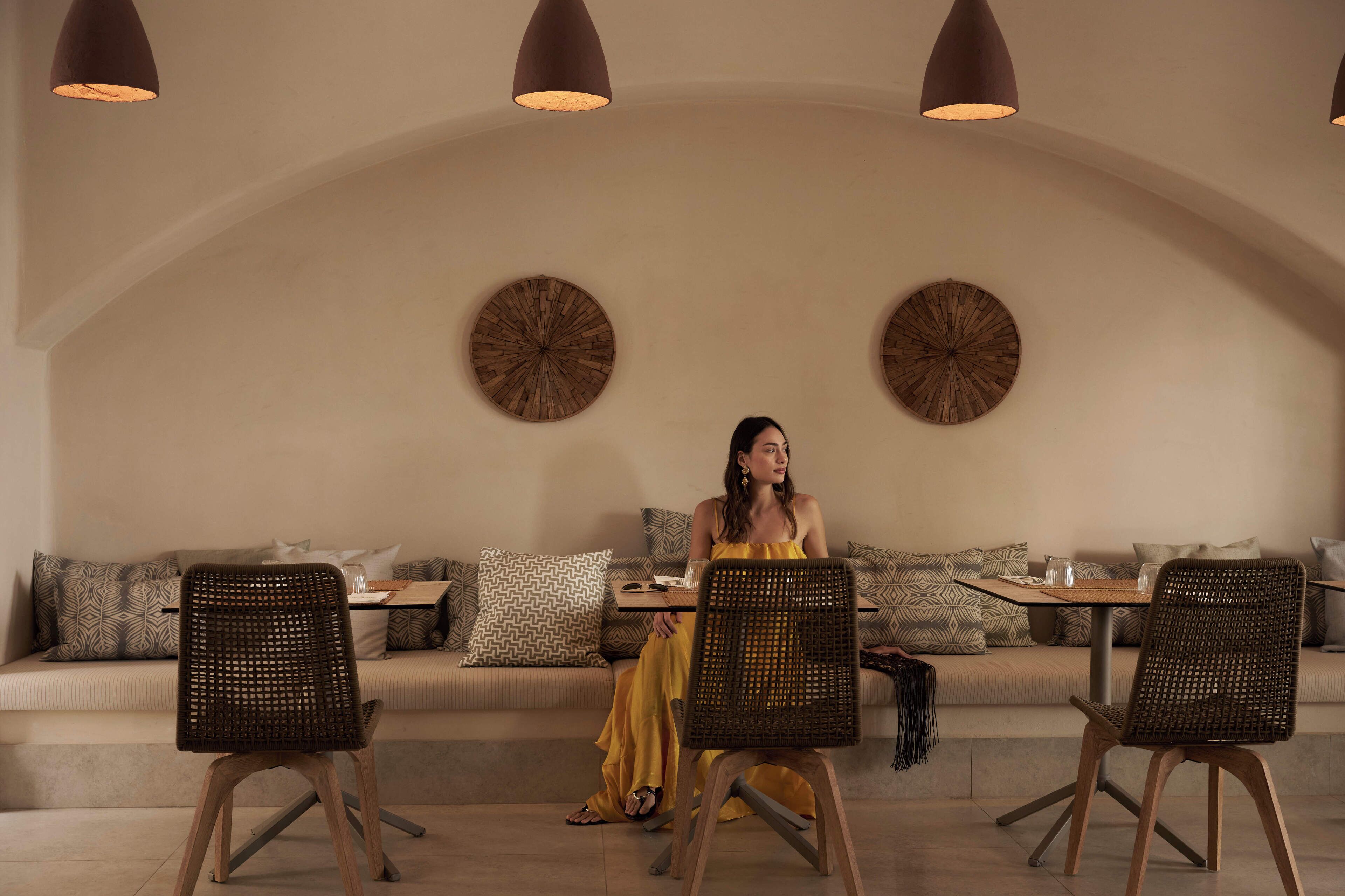 A woman seated at a table in a room with a high arched ceiling, patterned cushions, and two wall-mounted clocks.