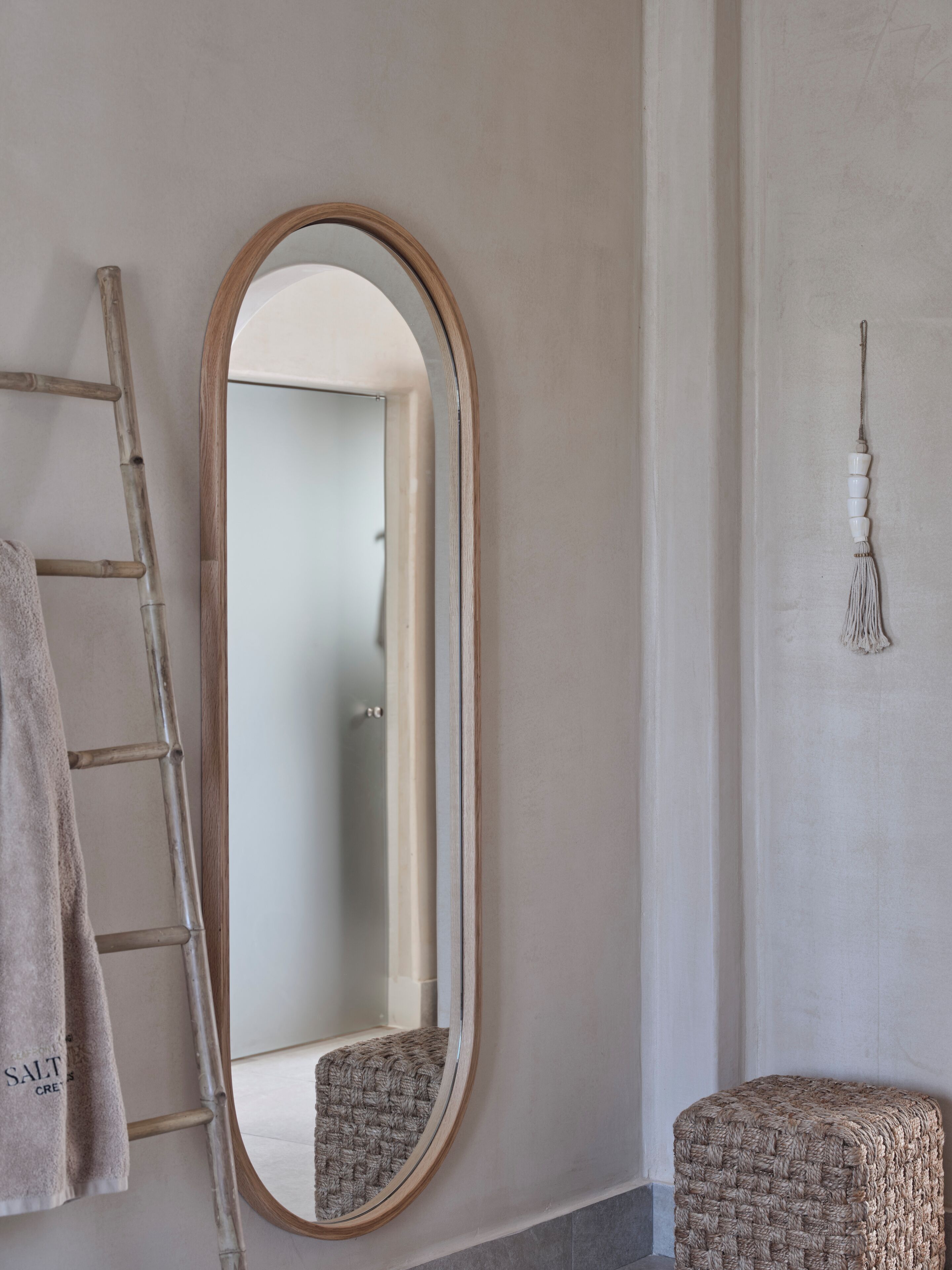 A bathroom with a wooden ladder, a round mirror, a woven stool, and a wall-mounted tassel.