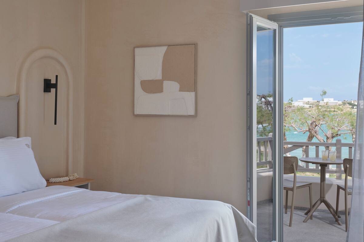 Quarto de hotel com cama, porta com puxador, obra de arte na parede e varanda com mesa e cadeiras.
