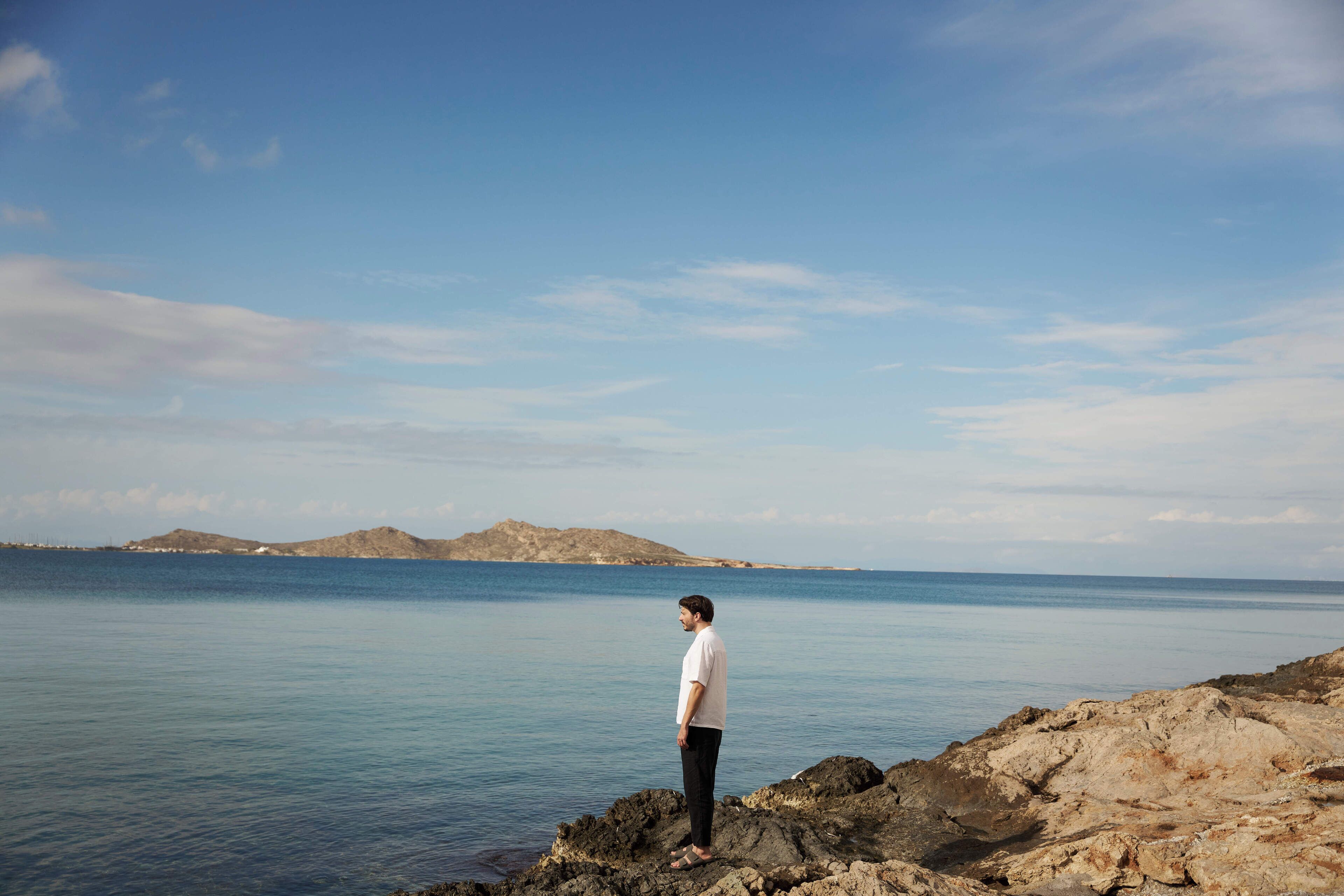 A person standing on rocky terrain by a calm body of water with distant landforms.