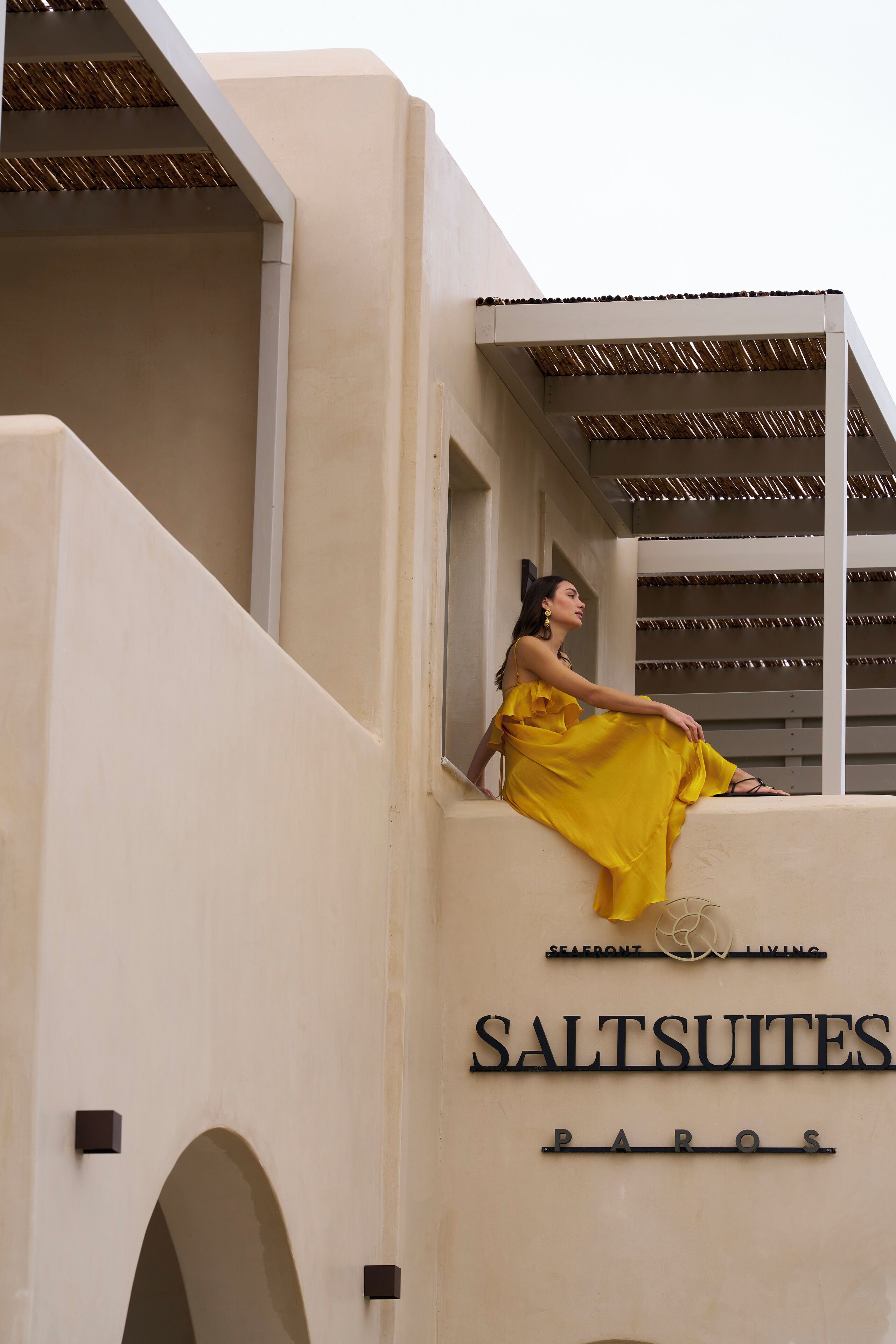 Une personne vêtue d’une robe jaune est assise sur le balcon d’un immeuble sur lequel on peut lire l’inscription « SALTSUITES PAROS ».