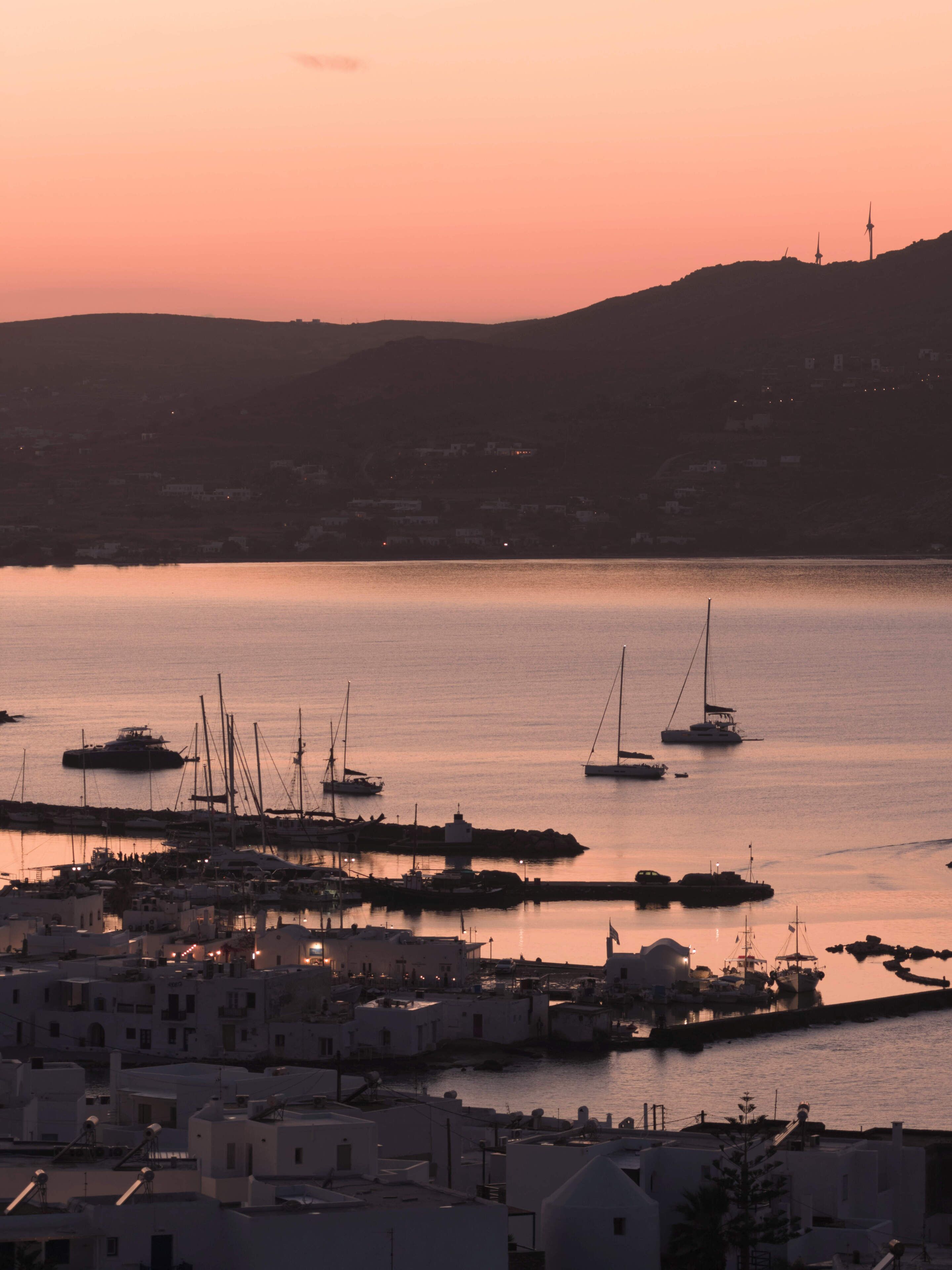 Un pueblo costero al atardecer con barcos amarrados en el puerto, edificios de techos planos y una colina al fondo.