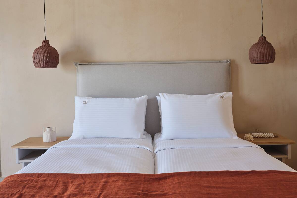 Cama bem arrumada com lençóis brancos e manta marrom, cabeceira cinza e duas mesas de cabeceira com um vaso branco e uma pequena planta.