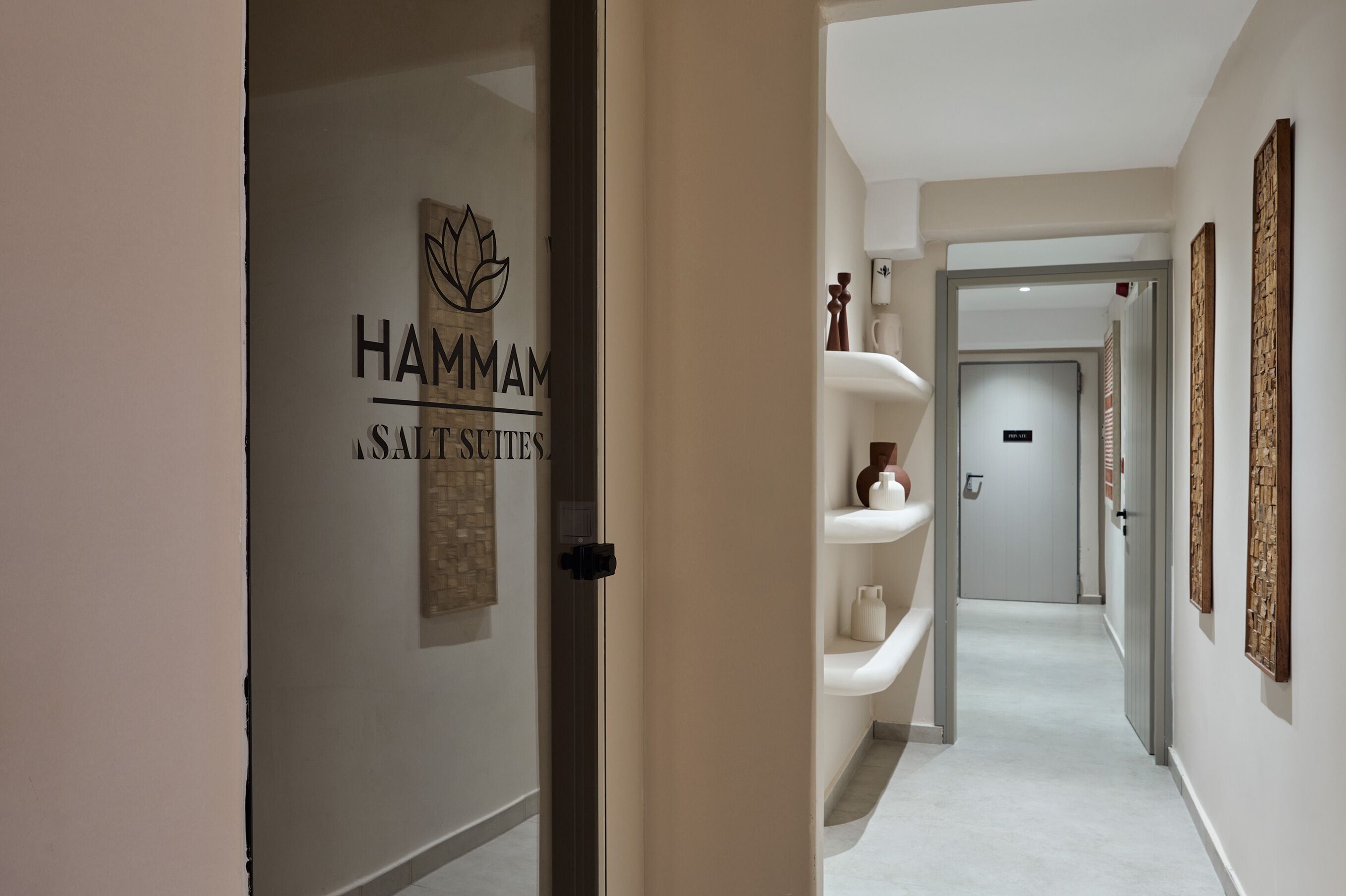 A hallway with a sign reading 'HAMMAM SALT SUITES,' a white wall, a door, and a shelf with decorative items.