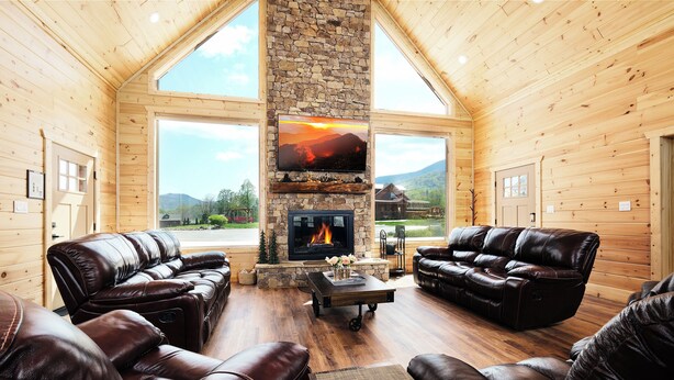 A log cabin interior with leather sofas, a stone fireplace, and large windows offering a mountain view. - Brand new gorgeous 5BR cabin with mtn views, game room, hot tub, movie theater! (Sevierville)