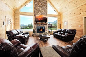 A log cabin interior with leather sofas, a stone fireplace, and large windows offering a mountain view. - Brand new gorgeous 5BR cabin with mtn views, game room, hot tub, movie theater! (Sevierville)