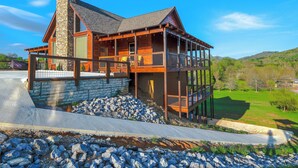 A modern house with a stone chimney, a balcony, and a view of a green landscape. - Brand new gorgeous 5BR cabin with mtn views, game room, hot tub, movie theater! (Sevierville)