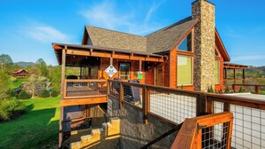 A multi-level wooden cabin with a stone chimney, wooden deck, and a view of the surrounding landscape. - Brand new gorgeous 5BR cabin with mtn views, game room, hot tub, movie theater! (Sevierville)