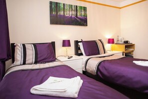 Camera d'albergo con due letti, entrambi con biancheria viola e cuscini a righe. Si vede un quadro raffigurante una foresta, un comodino con una lampada e un tavolino con una lampada e una macchina per il caffè.