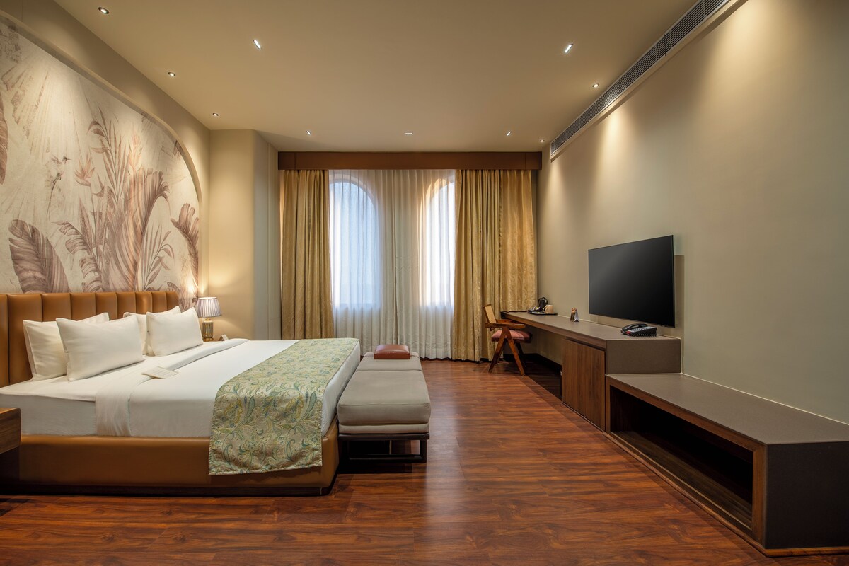 A modern hotel room with a large bed, a desk with a TV, and a large window with curtains.