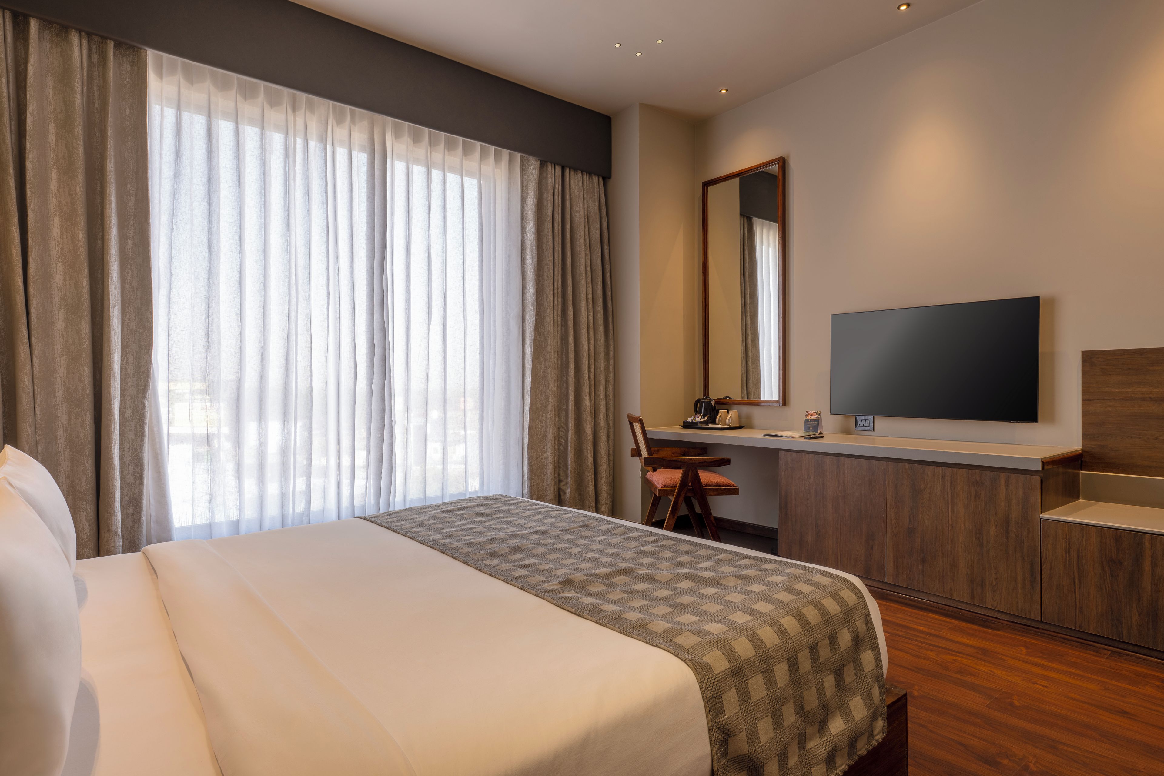 A hotel room with a large bed, a desk with a chair, a flat-screen TV, and a large window with curtains.