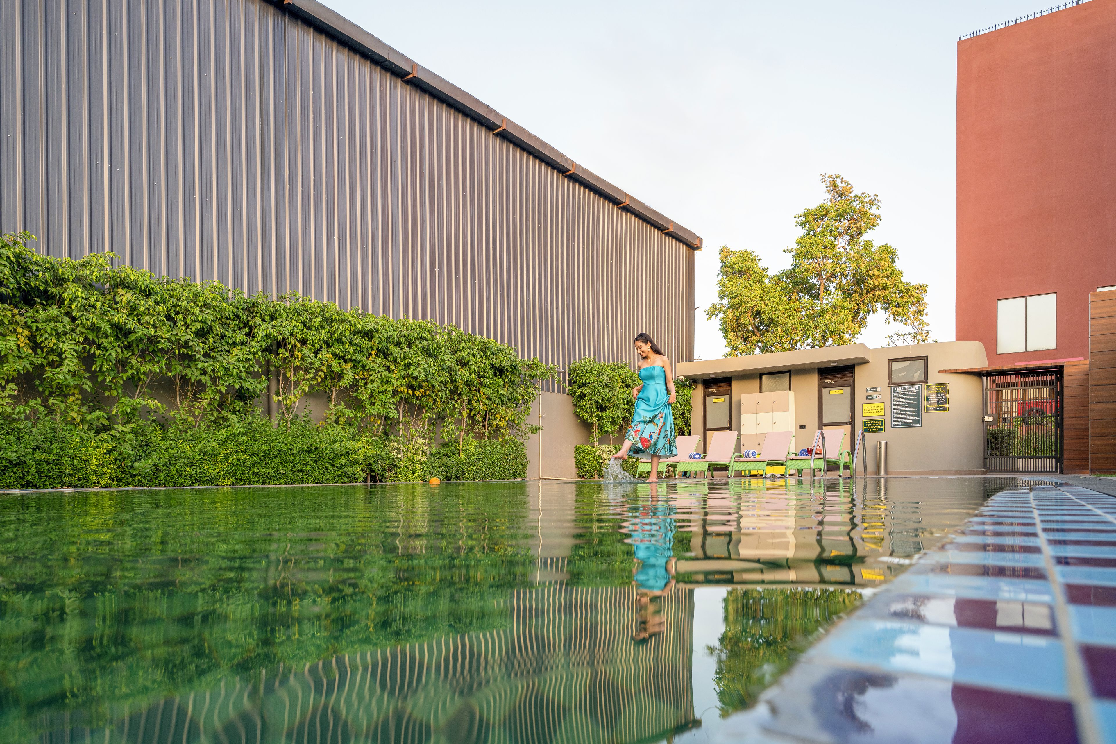 A swimming pool with a tiled edge, a person in a blue dress, and a building with a corrugated metal facade.