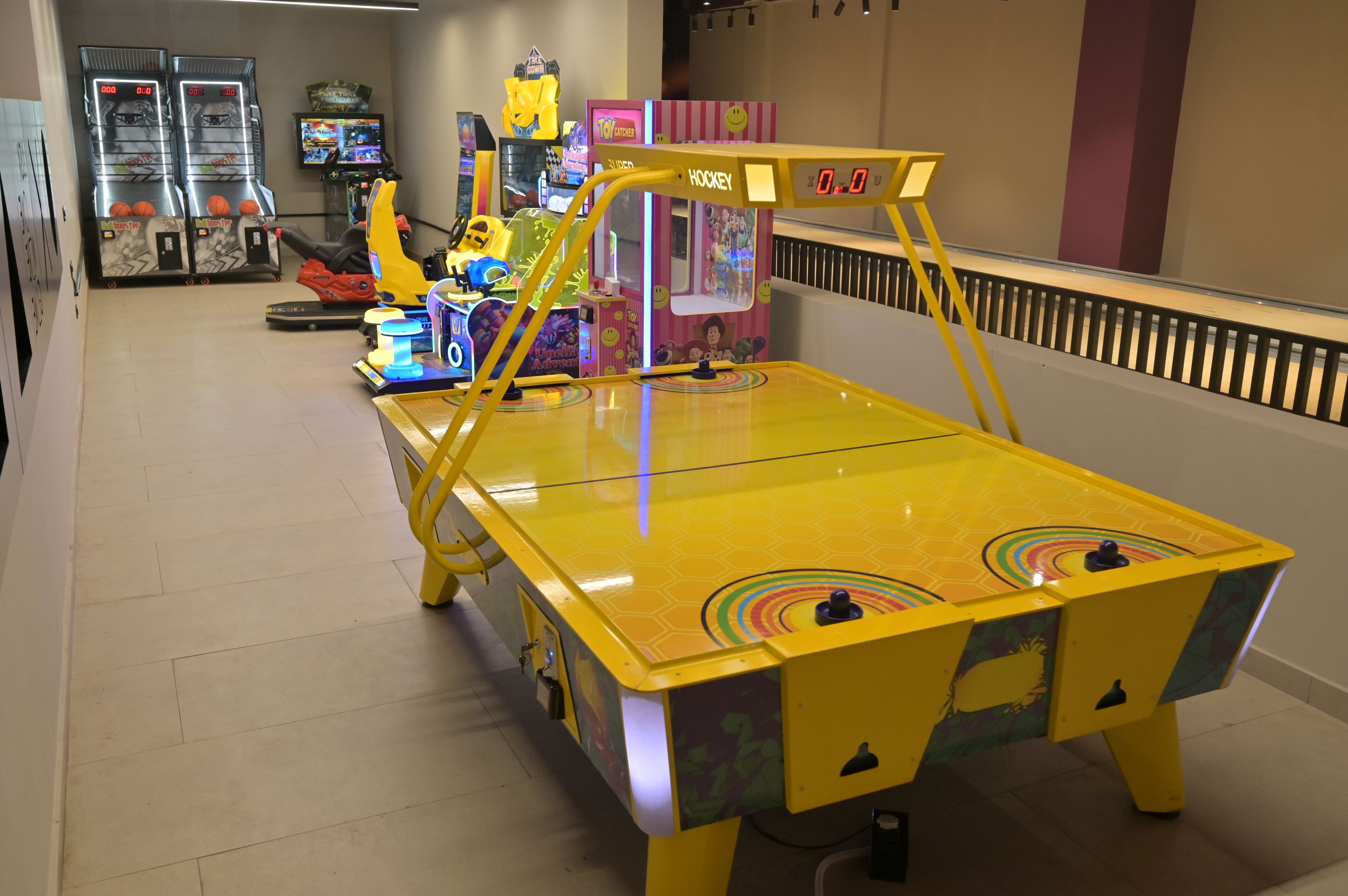 Um salão de jogos coberto, bem iluminado, com diversos jogos eletrônicos, incluindo uma mesa de pingue-pongue amarela e uma máquina de hóquei.